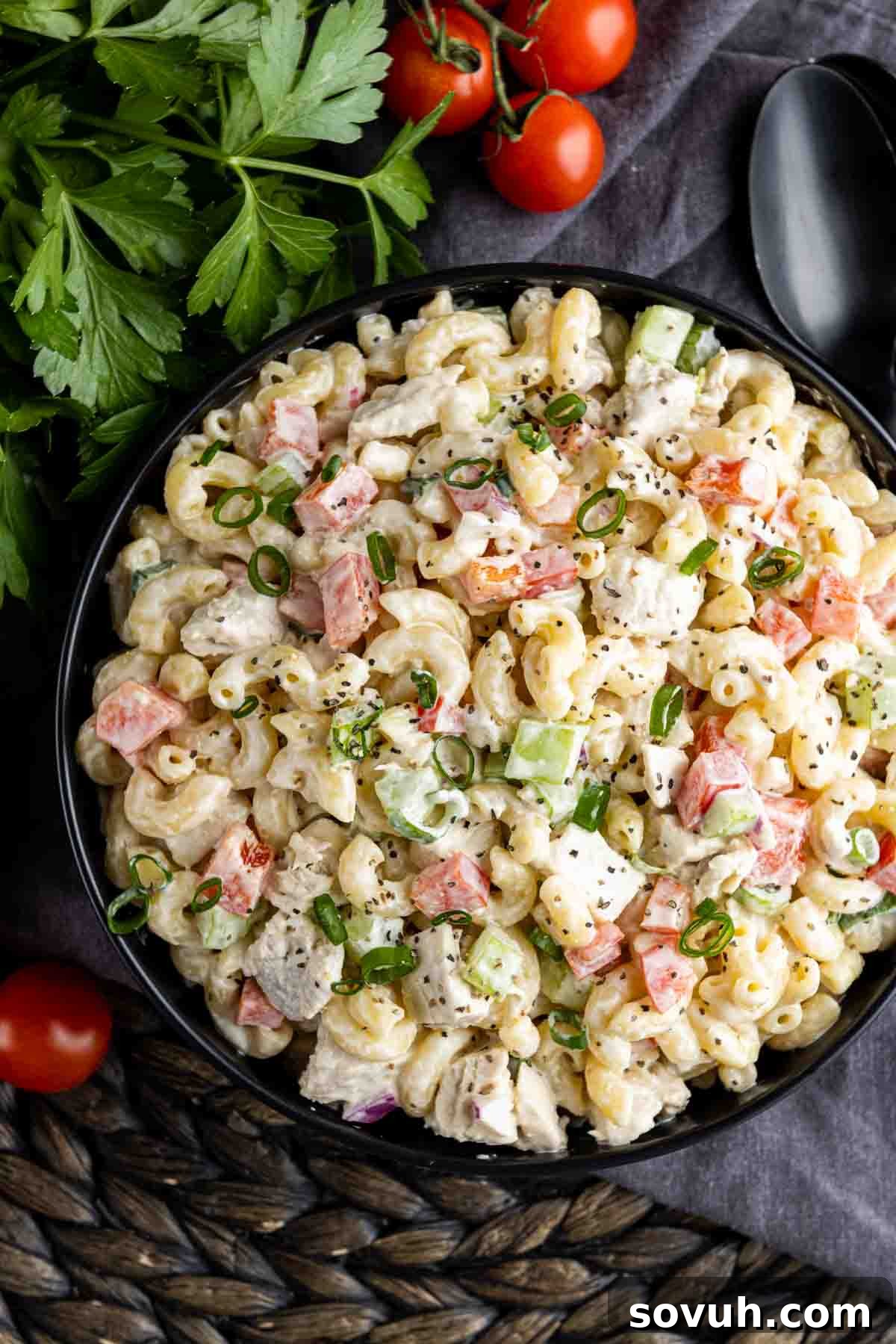 A black serving bowl filled with creamy Chicken Macaroni Salad, garnished with fresh parsley and a few cherry tomatoes on the side.
