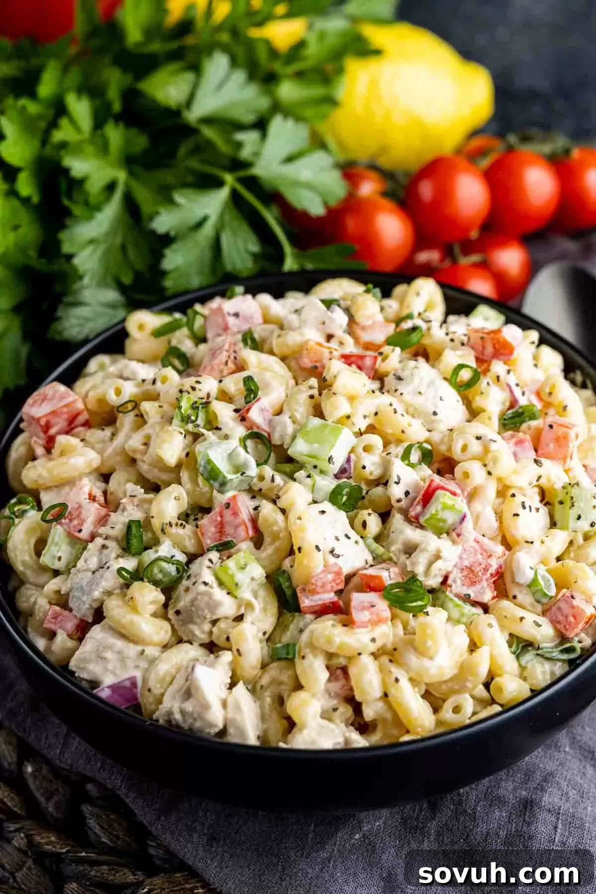A black bowl of creamy Chicken Macaroni Salad, garnished with fresh parsley, with cherry tomatoes and a lemon wedge in the soft-focus background.