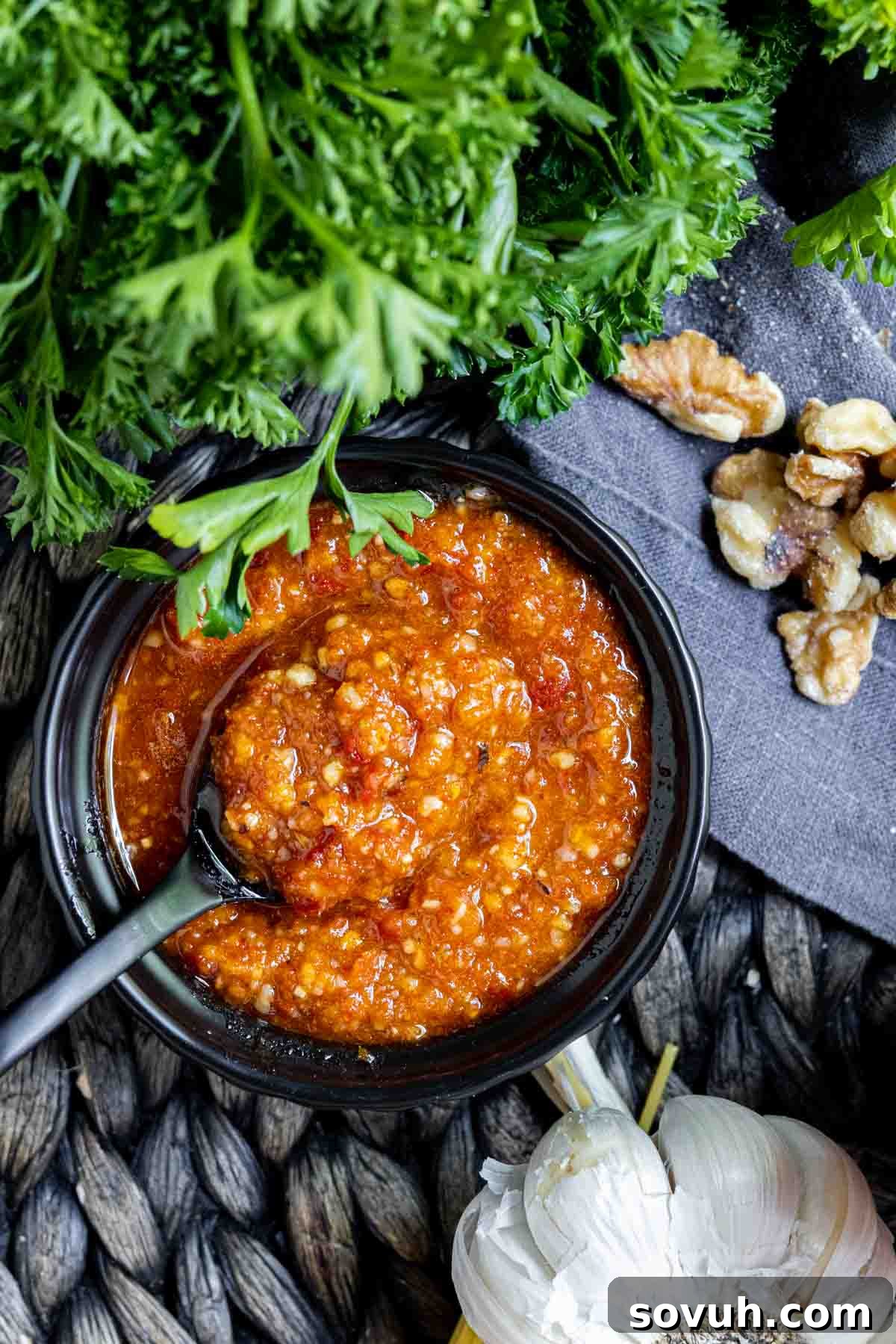 black spoon in a bowl of Pesto Rosso surrounded by parsley, walnuts and garlic