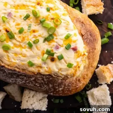 A loaf of French bread hollowed and filled with decadent creamy cheese and ham dip, topped with chopped green onions, surrounded by bread pieces on a rustic wooden surface.