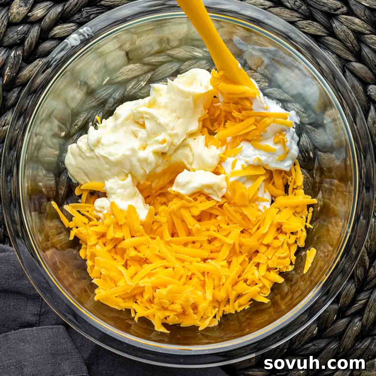 A bowl containing a creamy mixture of sour cream, mayonnaise, and shredded cheddar cheese, prepared for the topping of the John Wayne Casserole.