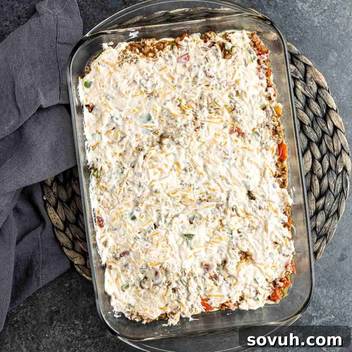 A John Wayne Casserole in a baking dish, now with the creamy sour cream and mayonnaise mixture spread smoothly over the ground beef layer.