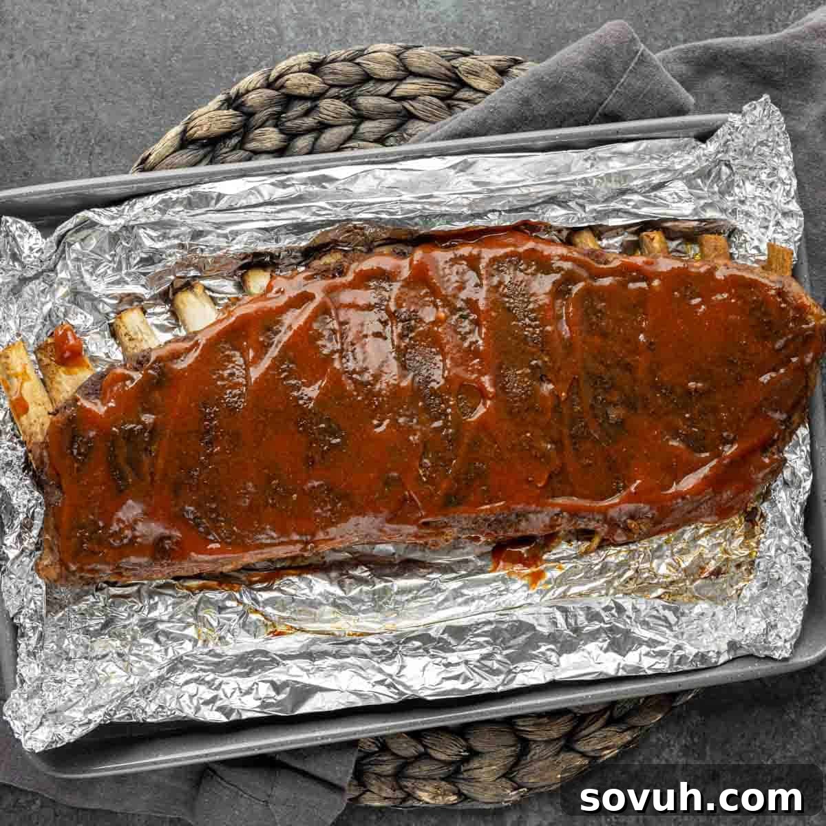 Overhead view of a rack of St Louis-Style Ribs covered in sauce, wrapped in foil on a tray with a rope handle.