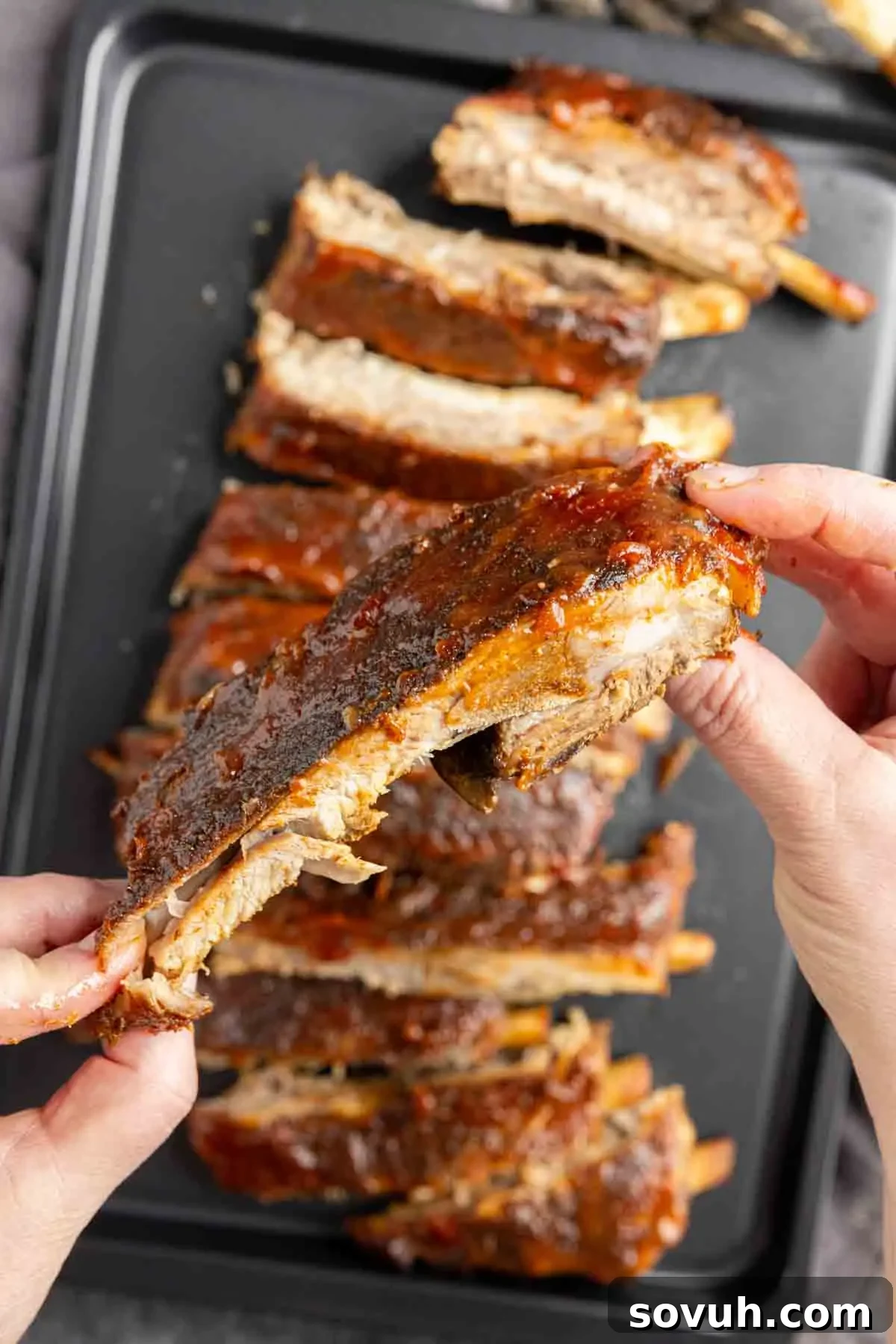 A person's hand pulling apart a tender, cooked St Louis-Style Ribs from a tray filled with barbecue ribs.