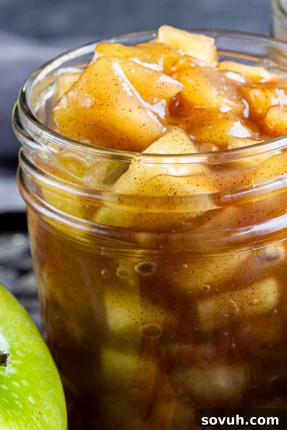 A jar of finished apple pie filling sits next to a slice of apple and cinnamon stick.