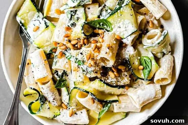 A bowl of pasta with creamy sauce, zucchini slices, fresh basil, and pine nuts, garnished with grated cheese and black pepper. A fork rests on the side of the bowl.