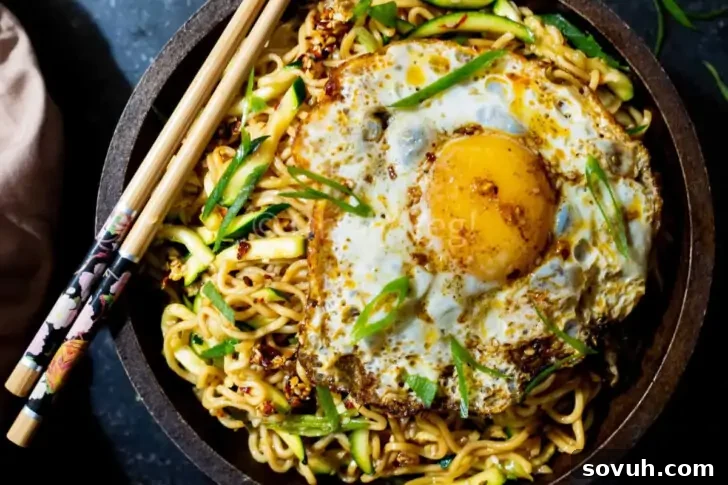 A bowl of stir-fried noodles topped with a fried egg, garnished with sliced green onions and served with chopsticks.