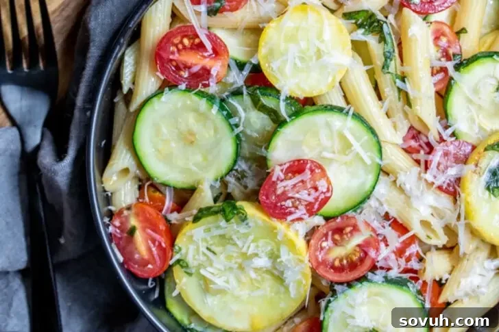Close-up of a bowl of pasta salad with penne, zucchini slices, cherry tomatoes, and grated cheese on top. A fork and napkin are placed beside the bowl.