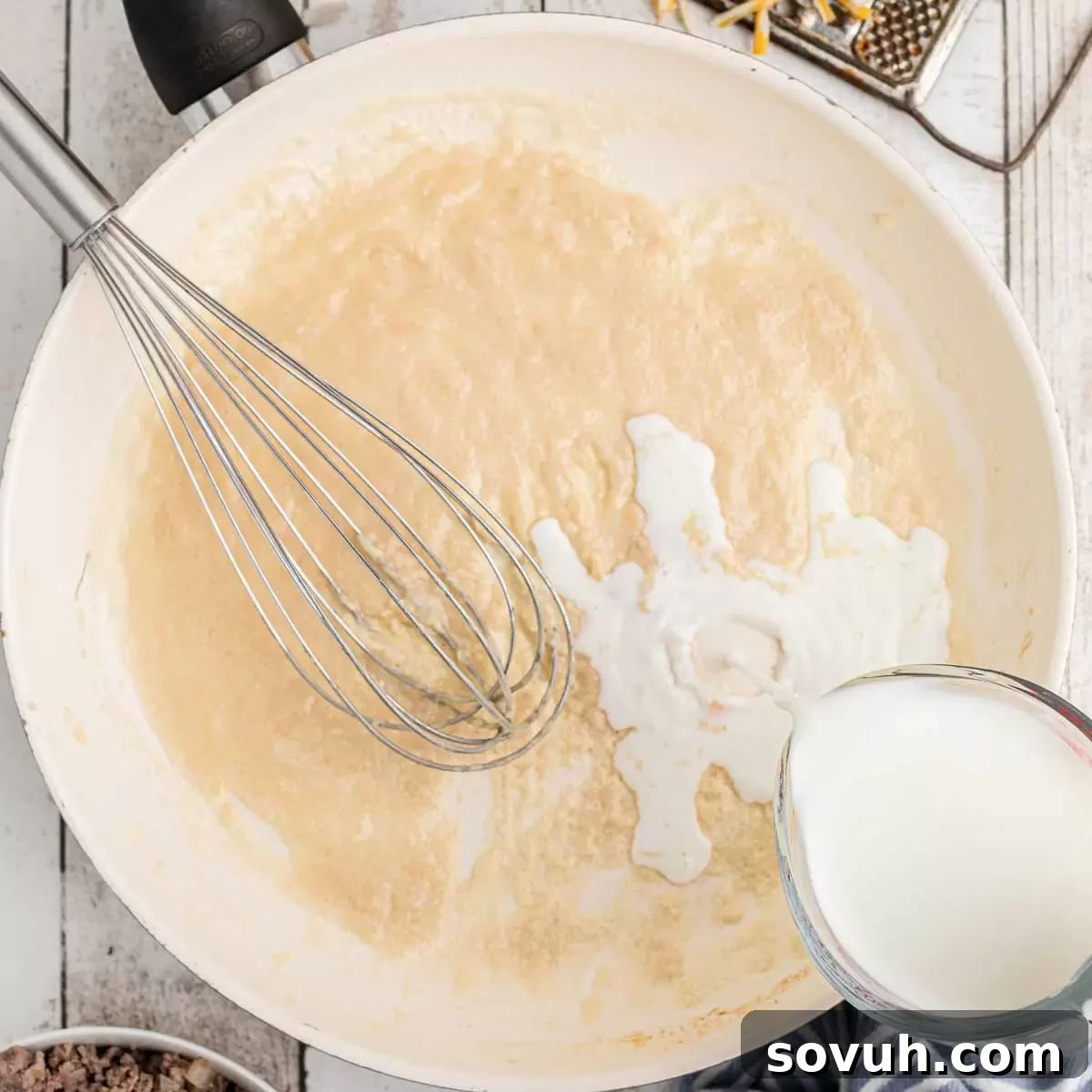 A skillet with a flour and butter roux, with warm milk being slowly poured in and whisked.