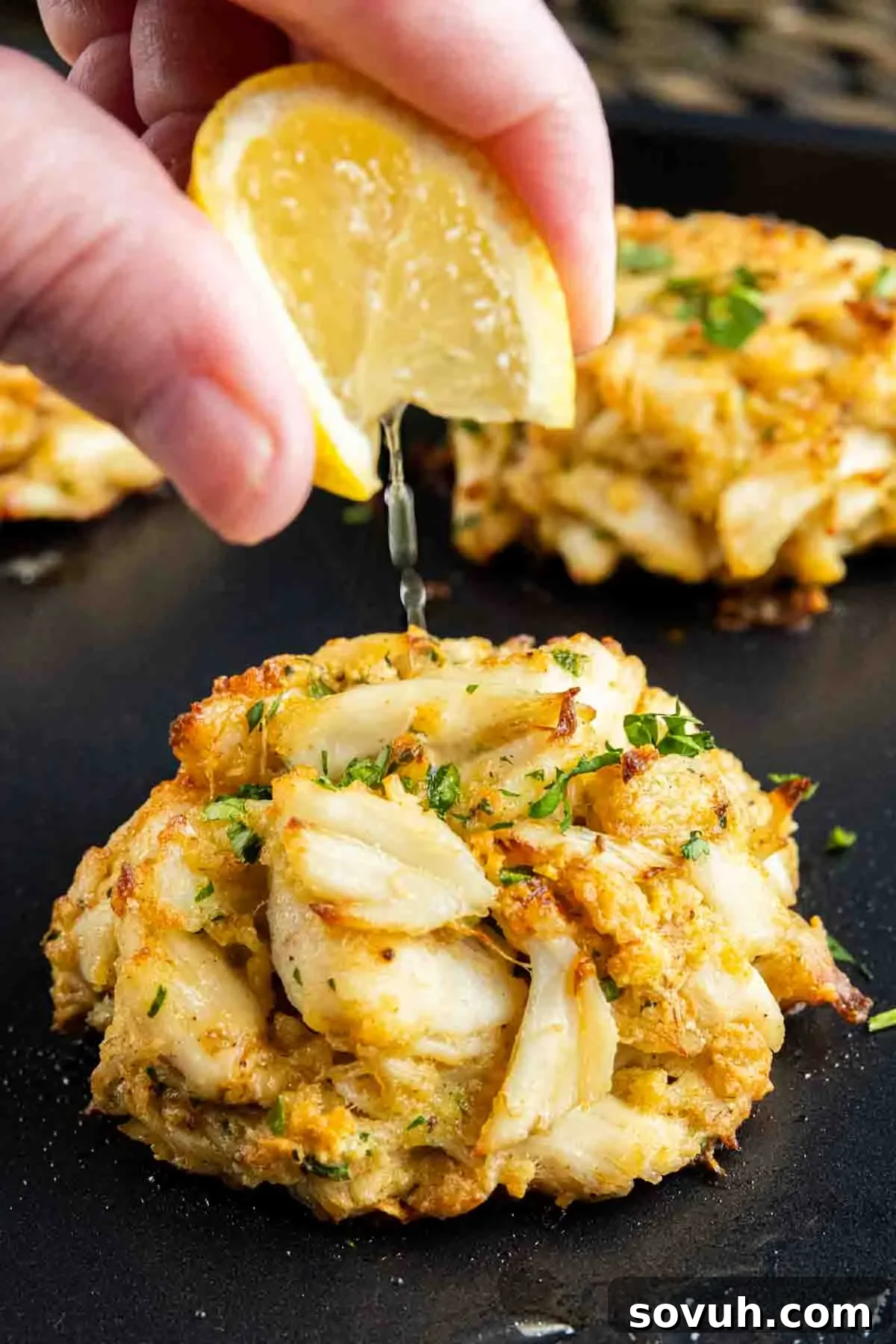 A person is drizzling lemon juice on some Maryland Crab Cakes.