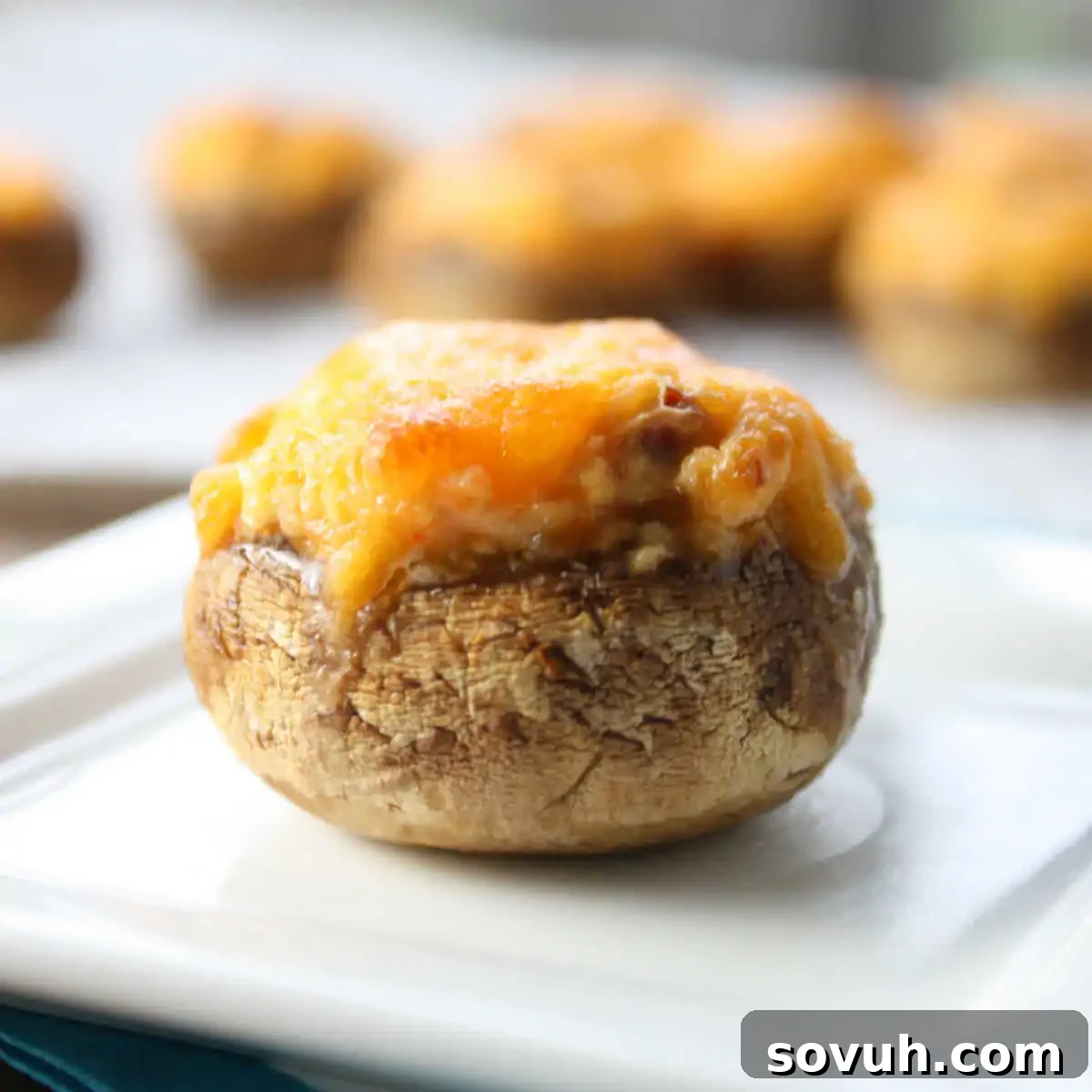 A close-up of a stuffed mushroom topped with melted cheese, placed on a square white plate with more stuffed mushrooms blurred in the background.
