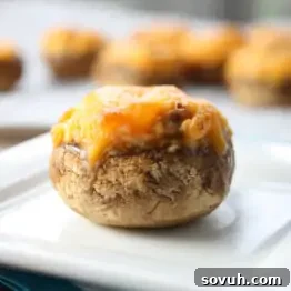 A close-up of a stuffed mushroom topped with melted cheese, placed on a square white plate with more stuffed mushrooms blurred in the background.