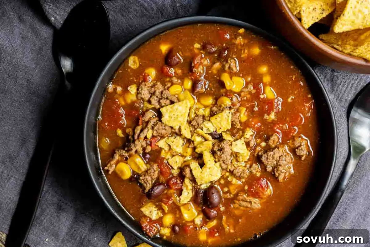 A rich bowl of taco soup, featuring savory ground meat, wholesome beans, sweet corn, and crunchy crushed tortilla chips, elegantly presented on a dark cloth background.