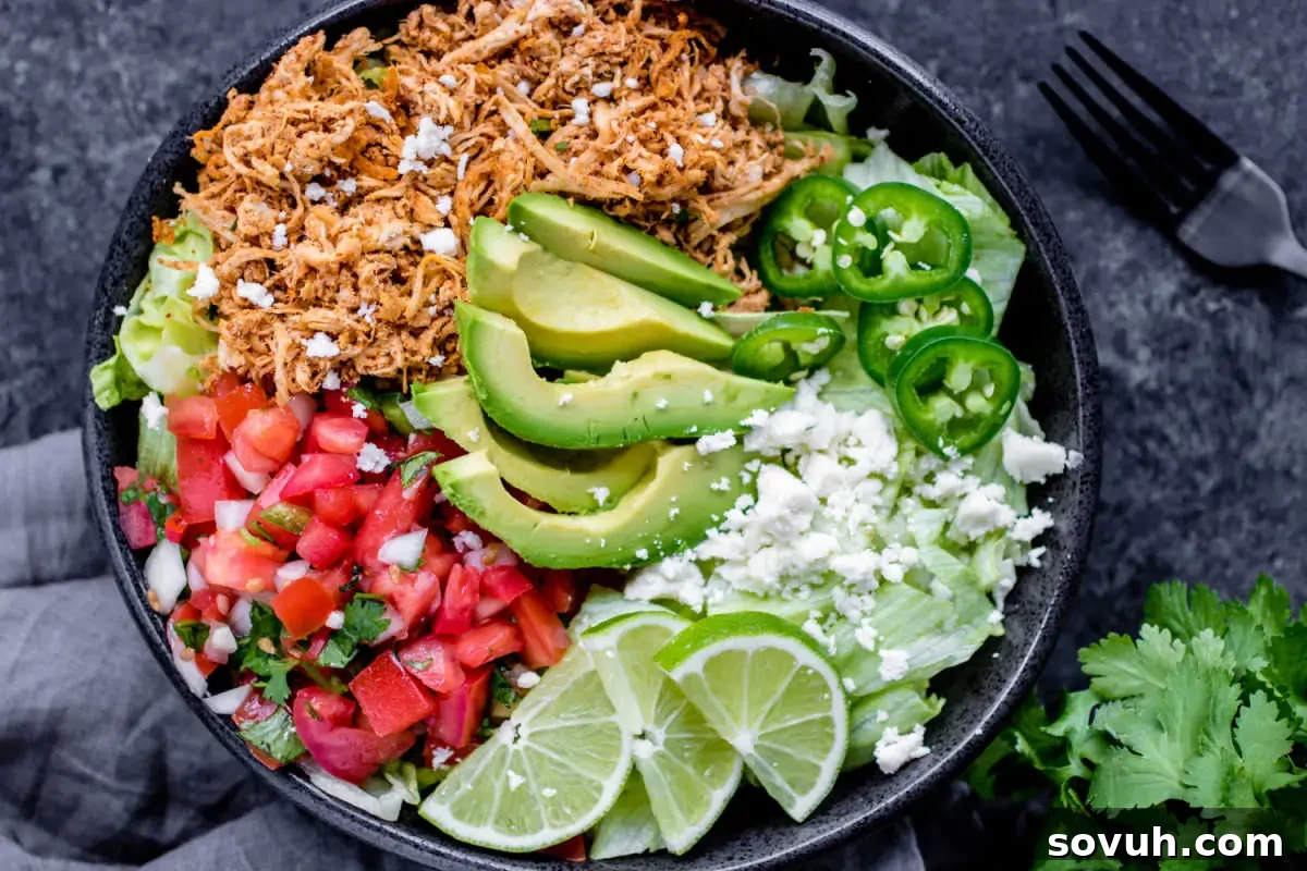 A vibrant and colorful taco salad bowl featuring shredded chicken, creamy avocado slices, fresh tomatoes, spicy jalapeños, crumbled cheese, and zesty lime wedges, all served in a stylish black bowl.
