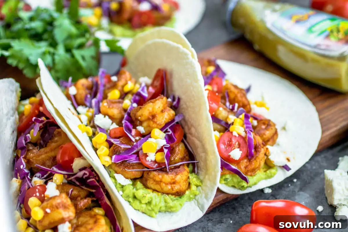 Three exquisite shrimp tacos, elegantly garnished with creamy guacamole, vibrant purple cabbage, sweet corn, and juicy tomatoes, beautifully presented with decorative flowers on a dark slate board.