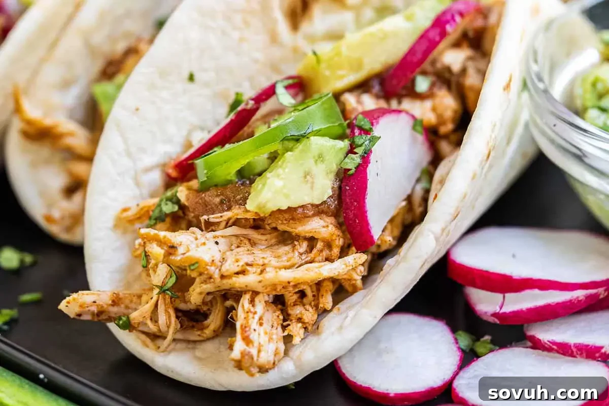 Three perfectly assembled chicken tacos topped with fresh avocado slices, vibrant radishes, and fragrant cilantro, served on a sleek black plate.