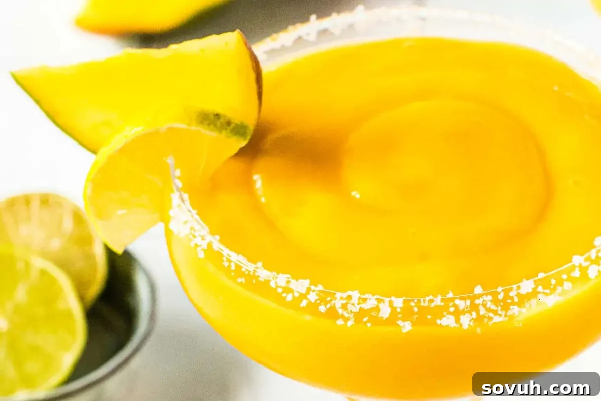 A close-up of a mango margarita garnished with a lime wedge and salt on the rim, with additional lime slices in the background.