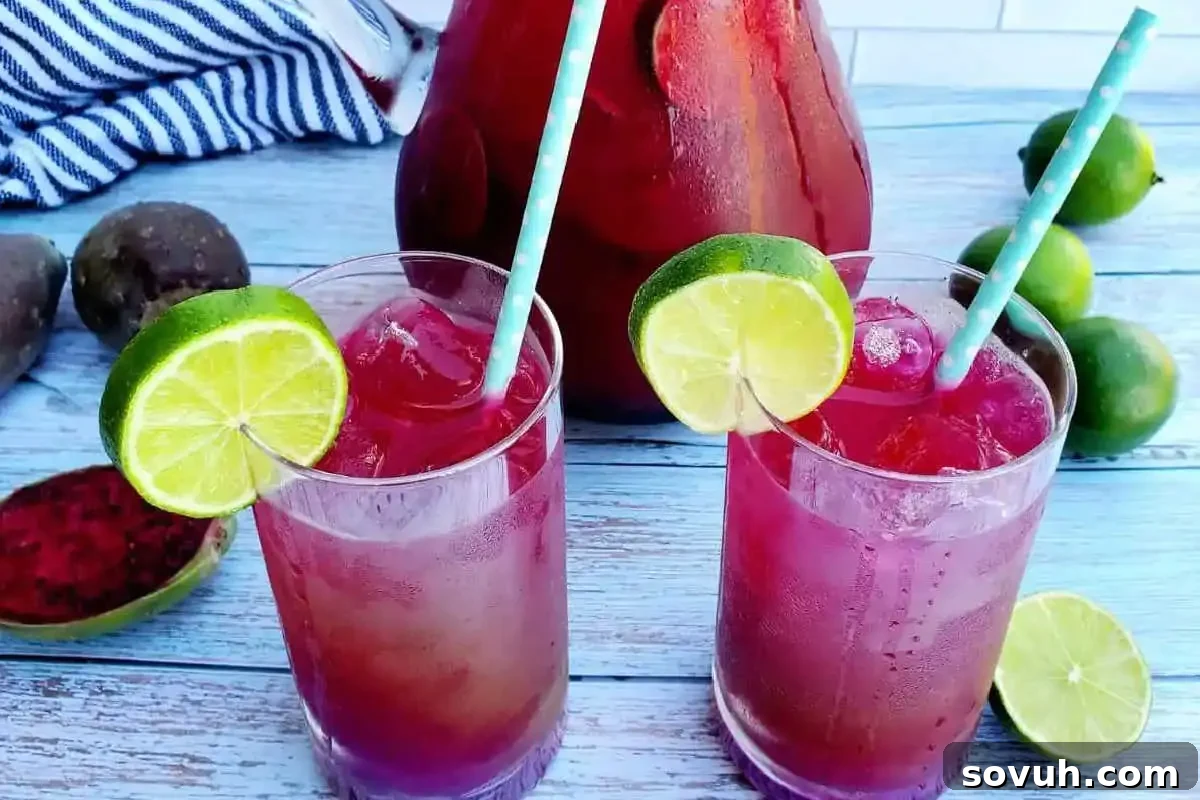 Two glasses of red iced drink with lime slices and straws on a wooden table, surrounded by fruits and a pitcher.