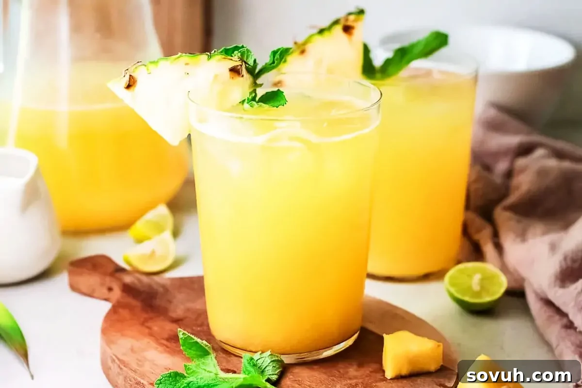 Two glasses of pineapple juice with mint garnish, lime wedges, and pineapple slices on a wooden board.
