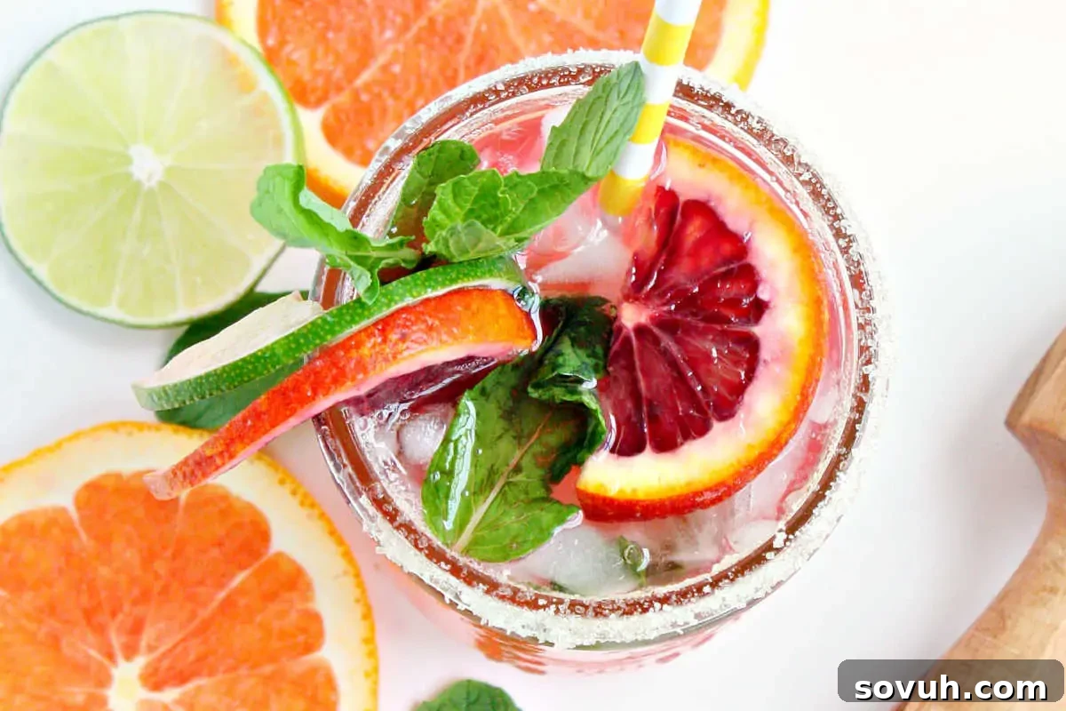 A refreshing cocktail garnished with a slice of blood orange, lime, and mint, served in a glass with a sugared rim and a striped straw.