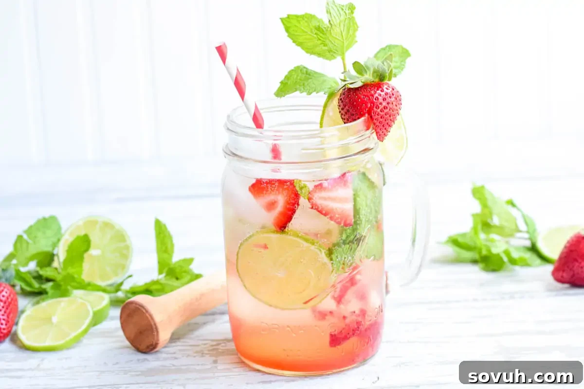 A refreshing drink in a mason jar with slices of lime, strawberry pieces, a mint garnish, and a striped straw, set against a white wooden background.