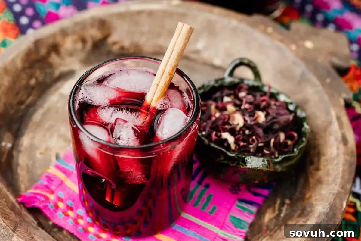A glass of iced hibiscus tea with a cinnamon stick, served on a colorful woven cloth beside a bowl of dried hibiscus flowers.