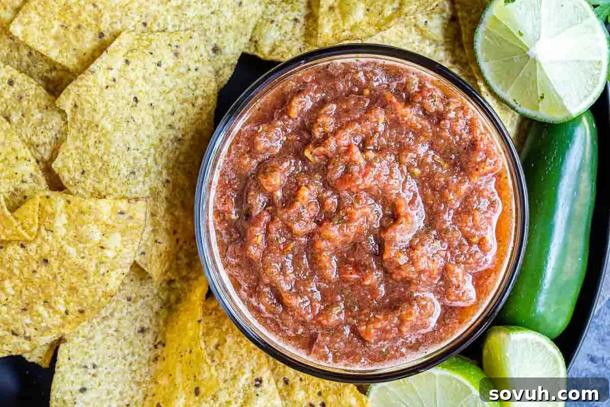 A large bowl of vibrant red salsa, made with diced tomatoes, surrounded by golden tortilla chips, fresh lime wedges, and a whole zucchini on a sleek black surface, ready for a Cinco de Mayo celebration.