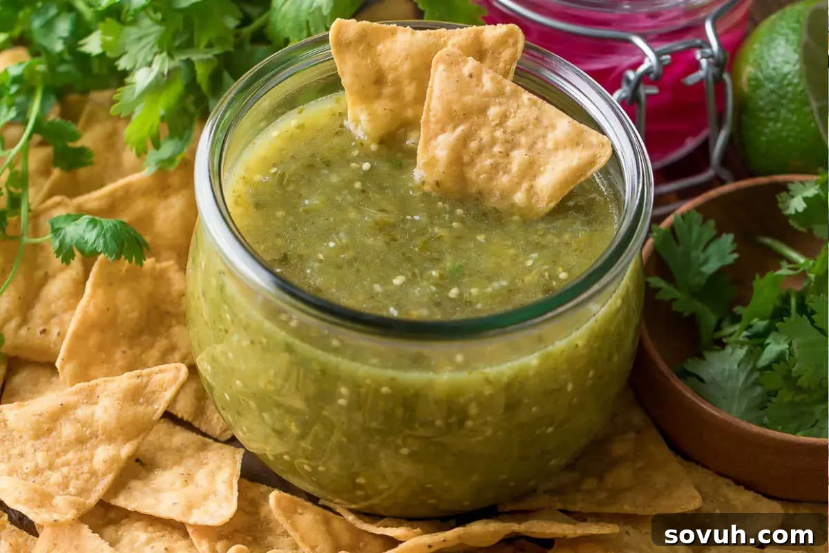 A clear glass jar filled with vibrant green salsa verde, perfectly accompanied by a scattering of golden tortilla chips and fresh cilantro leaves, all set on a rustic wooden surface.