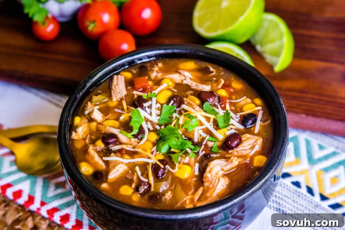 A bowl of chicken tortilla soup with beans and corn, garnished with shredded cheese and cilantro, accompanied by lime wedges and cherry tomatoes.