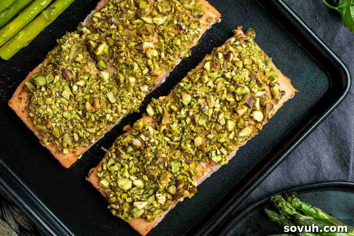 Three pistachio-crusted salmon fillets on a black baking tray, served with asparagus, on a wooden table.