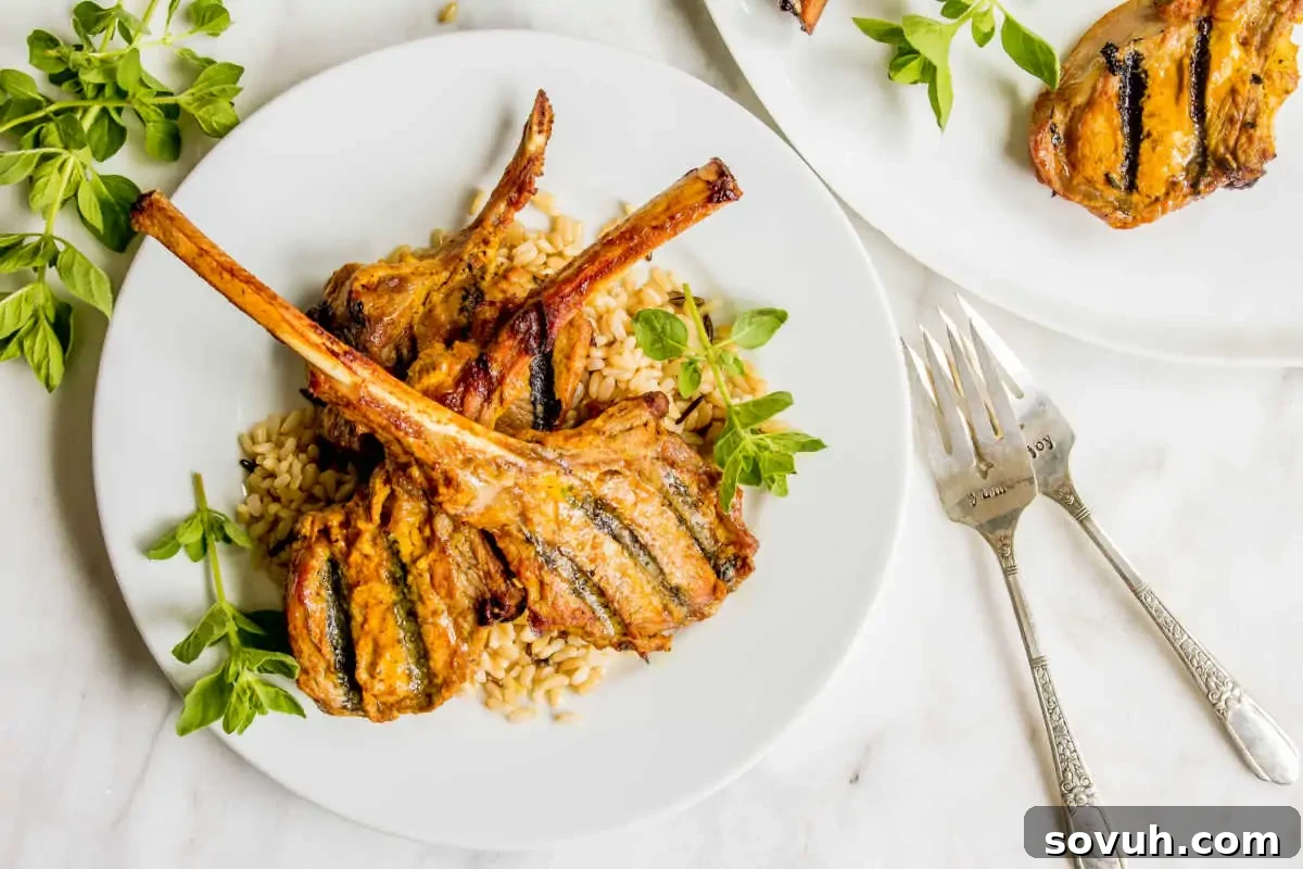 Grilled lamb chops served with barley and garnished with basil on white plates, accompanied by silver forks on a marble surface.