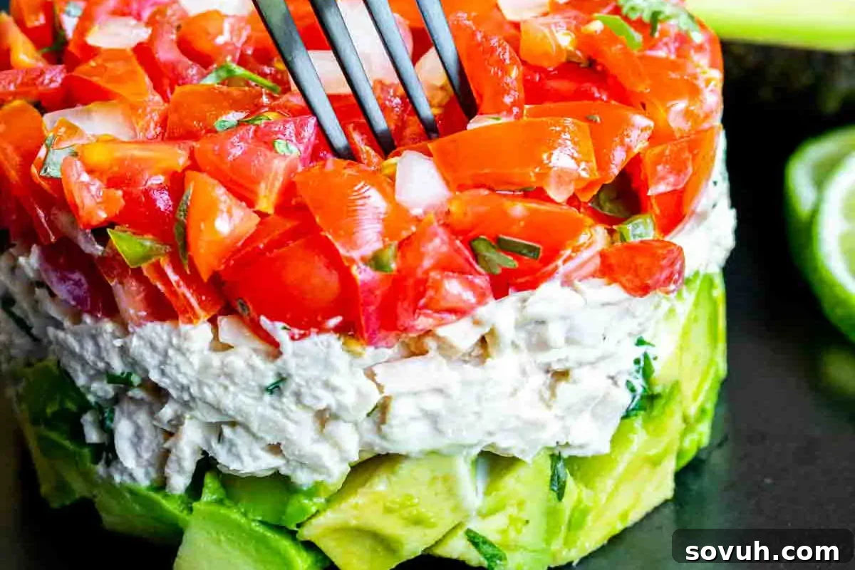 A vibrant avocado salad stack with layers of chopped avocado, seasoned cream cheese, and diced tomato, topped with fresh herbs and a fork.