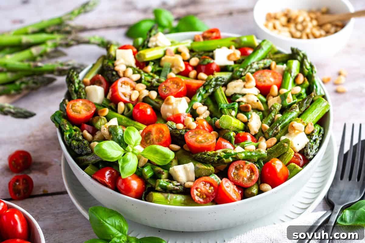 A vibrant salad with asparagus, cherry tomatoes, pine nuts, and basil in a white bowl on a wooden table.