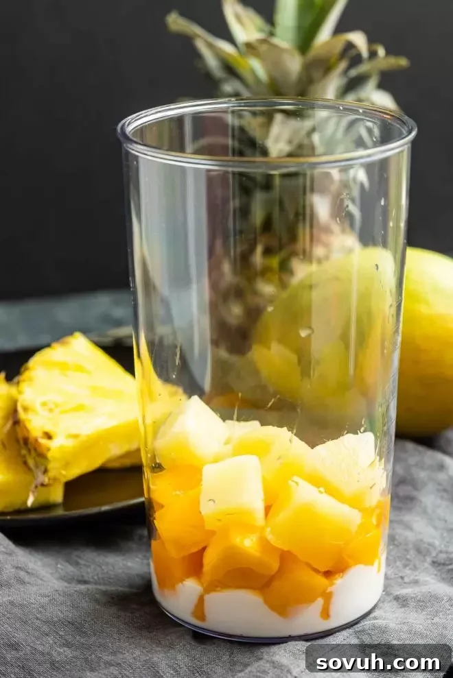 A blender being loaded with vibrant mango, fresh pineapple, a splash of almond milk, and creamy Greek yogurt, ready to be blended into a delicious smoothie.