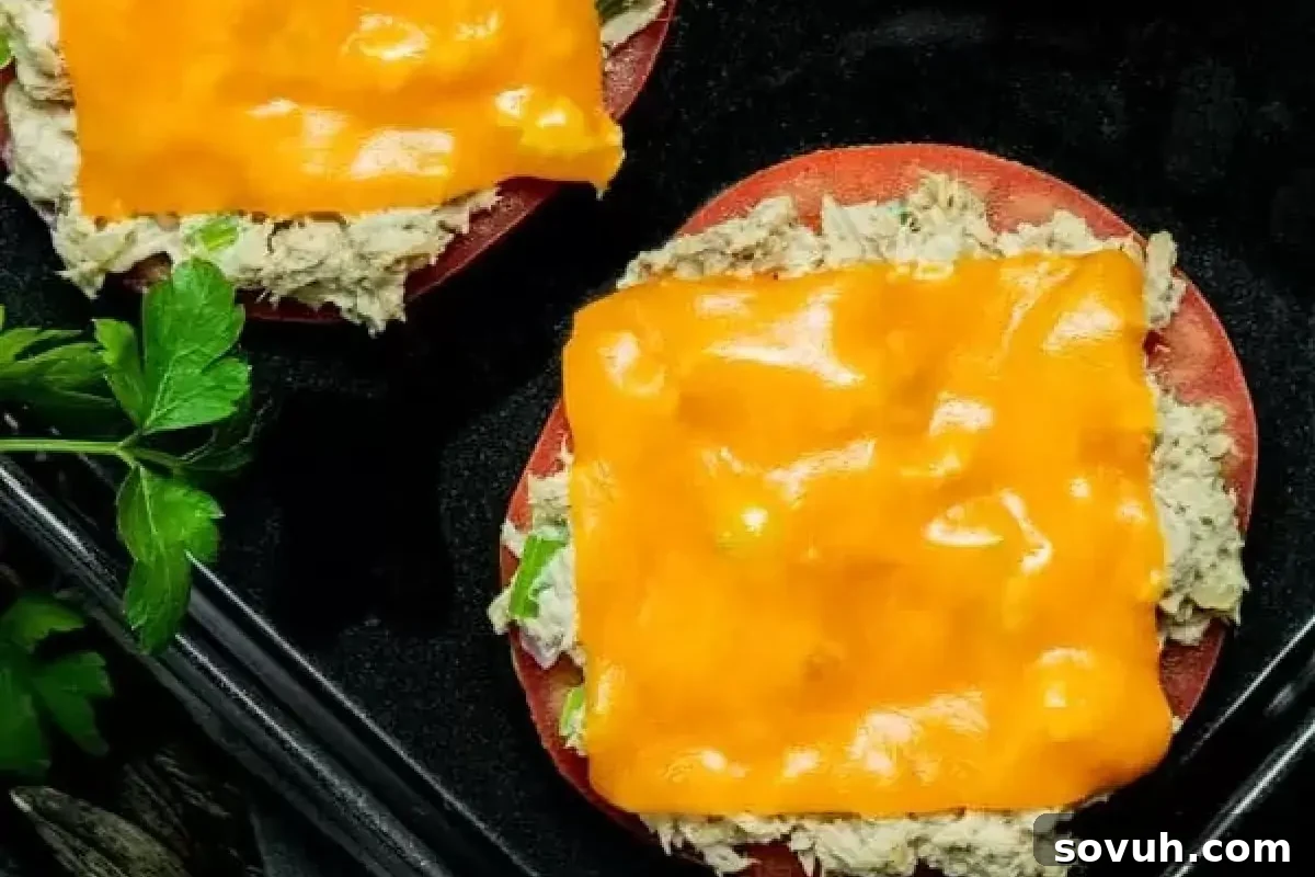 Two open-faced tuna melts with melted cheddar cheese on tomato slices, served on a black plate garnished with parsley.