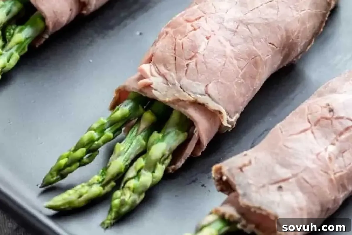 Slices of roast beef rolled around fresh asparagus spears on a dark plate.