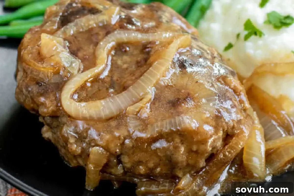 Classic Homemade Salisbury Steak on a dark plate, featuring tender ground beef patties smothered in a rich onion gravy, perfectly complemented by creamy mashed potatoes and vibrant green beans.