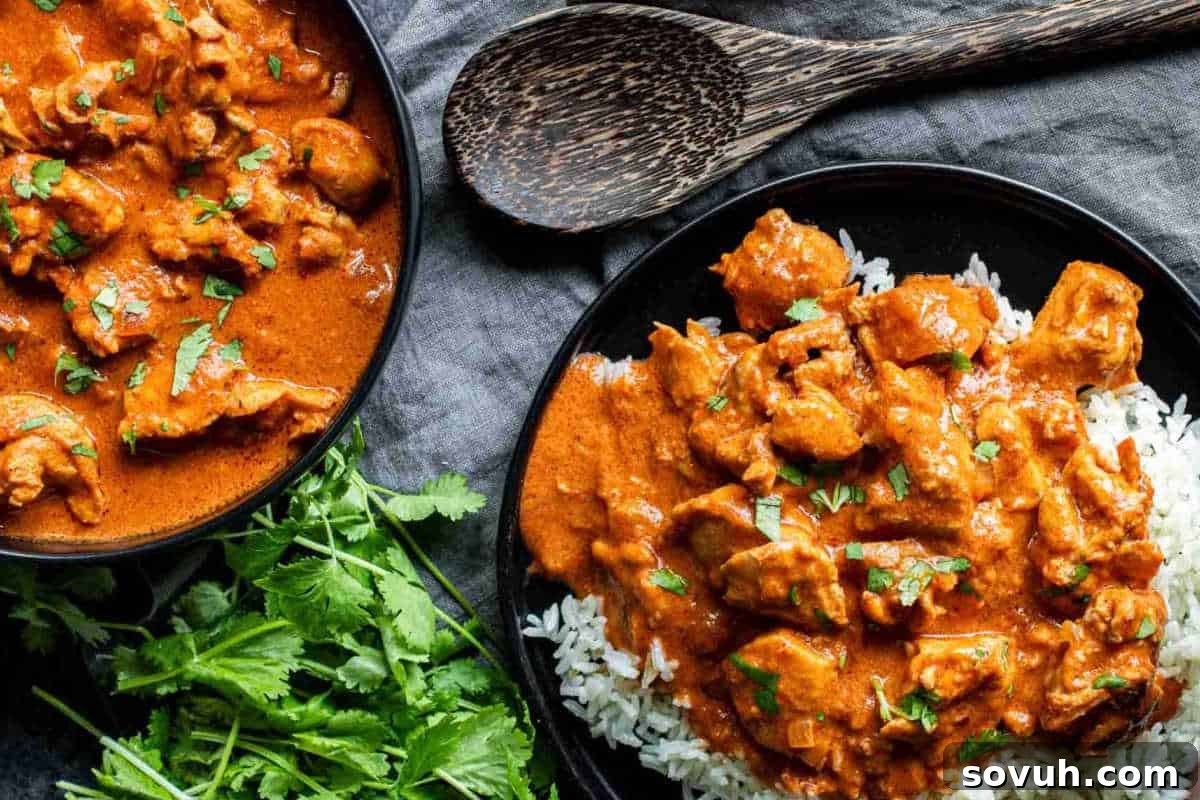 A plate of chicken tikka masala served over rice, garnished with herbs, next to a wooden spoon and fresh cilantro on a dark textured surface.