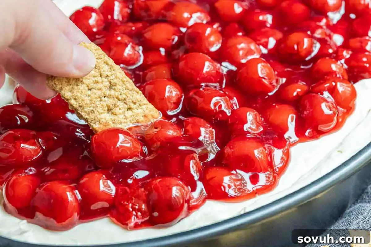 A hand dipping a graham cracker into a cherry pie with a rich, glossy filling and cream cheese base.