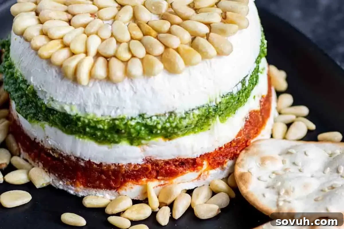 A layered cheese and pesto terrine with pine nuts on top, served with crackers on a black slate board.