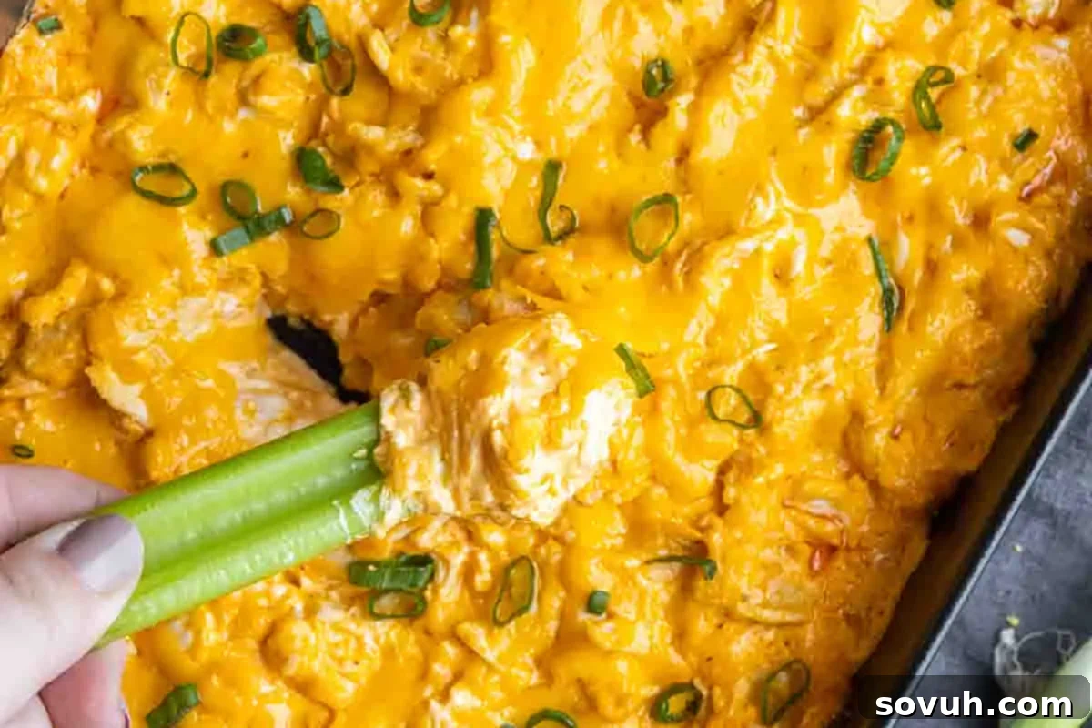 Close-up of buffalo chicken dip in a baking dish, garnished with green onions, served with a celery stick being dipped in.