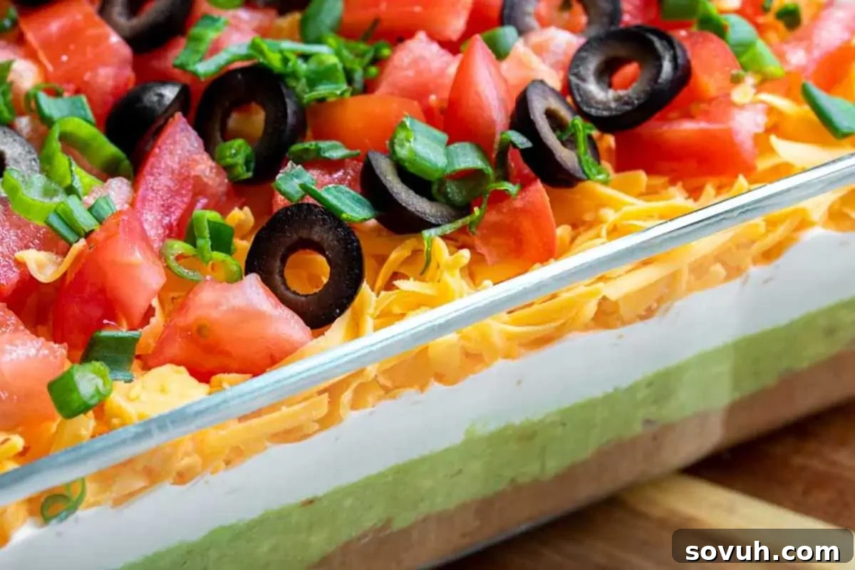 A close-up view of a seven-layer dip in a glass dish, showcasing layers of beans, guacamole, sour cream, shredded cheese, diced tomatoes, sliced black olives, and chopped green onions—perfect for your favorite dunk-worthy dip recipes.