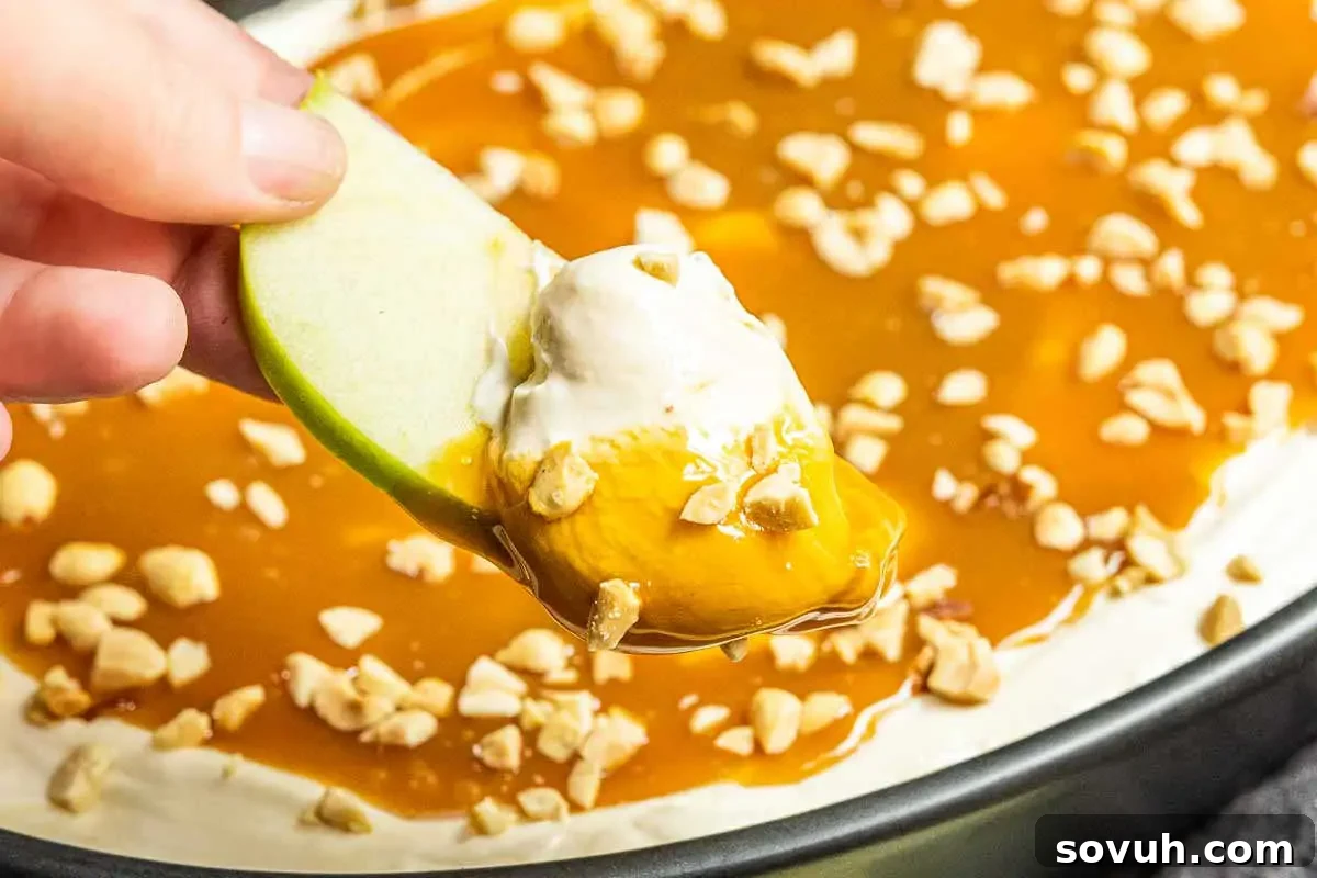 A hand dipping a green apple slice into caramel sauce topped with whipped cream and crushed peanuts.