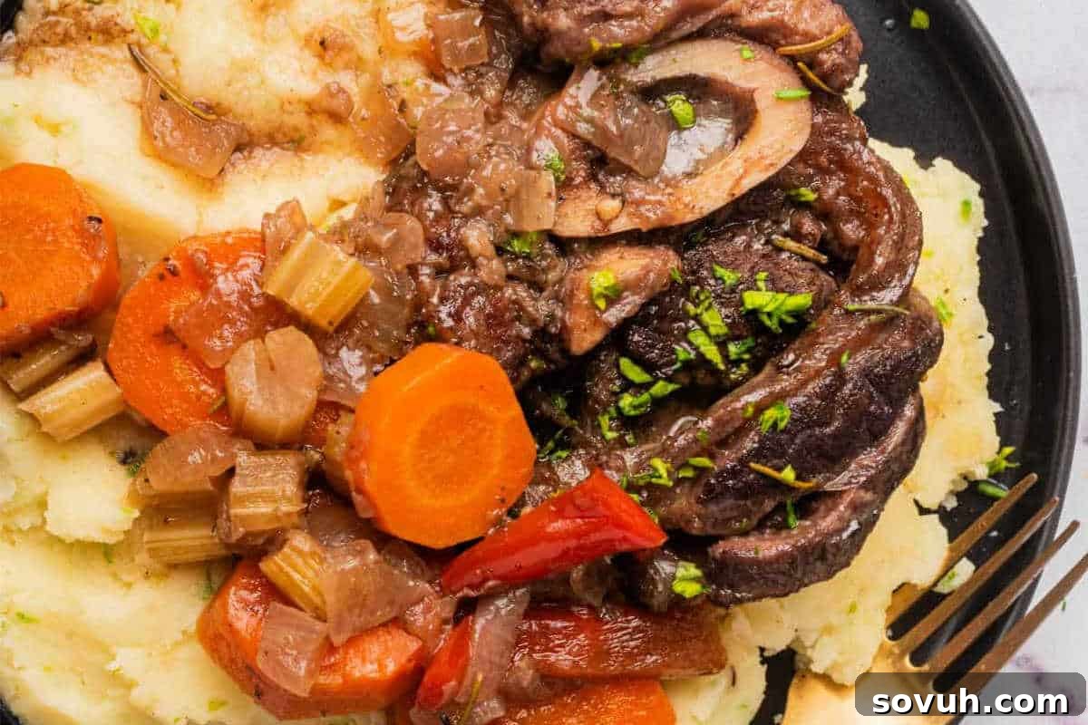 A plate of braised meat served with carrots, celery, and onions on top of mashed potatoes, garnished with chopped herbs.