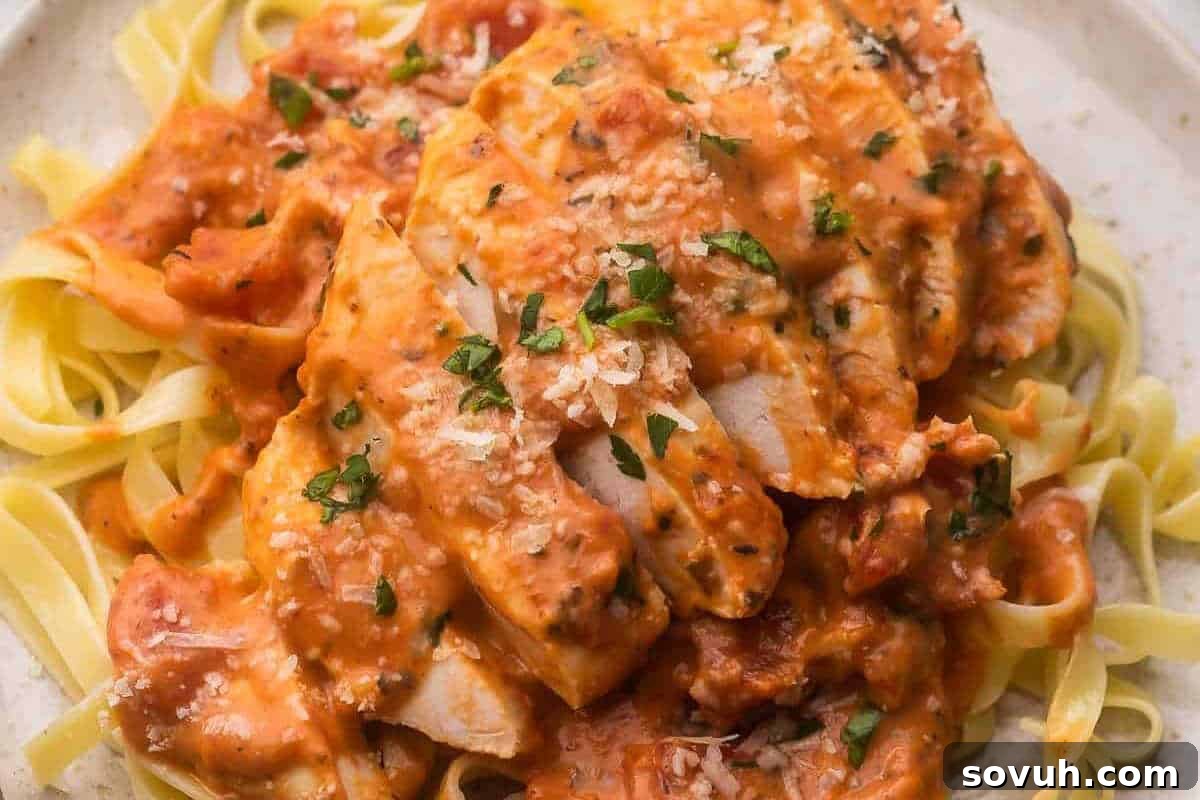 A plate of fettuccine pasta topped with sliced chicken breast in a creamy tomato sauce, garnished with chopped parsley and grated Parmesan cheese.