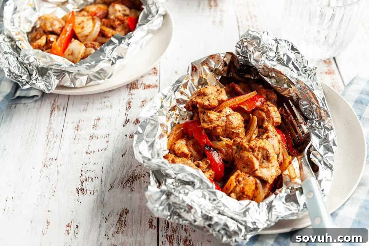 Two generous servings of perfectly cooked chicken and colorful vegetables, vibrant with Cajun seasoning, neatly contained in individual aluminum foil packets and arranged on white plates atop a rustic wooden table.