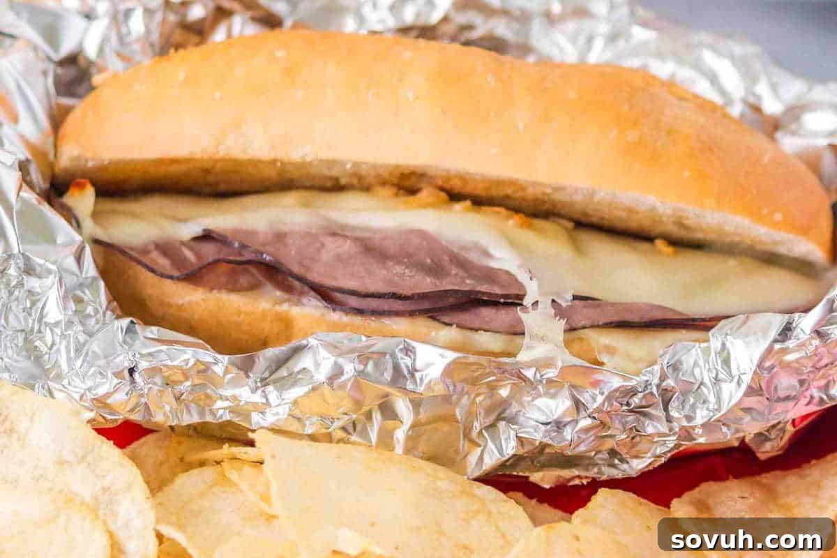 A savory roast beef sandwich, topped with gooey melted cheese, nestled in a sub roll and partially wrapped in aluminum foil, served alongside crispy potato chips.