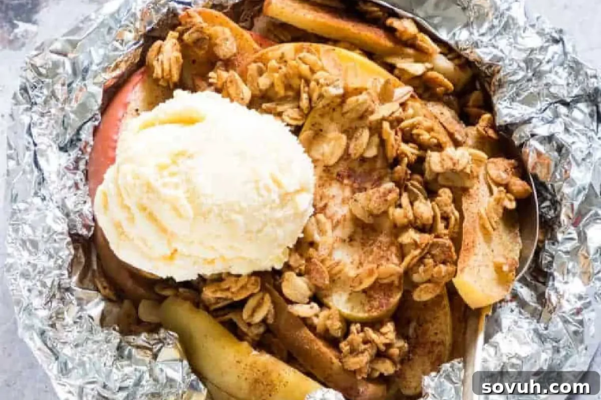 A comforting dessert featuring perfectly baked apple slices mingled with a crunchy oat topping, generously crowned with a scoop of melting vanilla ice cream, all nestled within a foil packet.