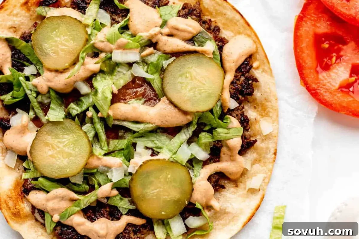 Open-faced taco with ground meat, diced onions, shredded lettuce, sliced pickles, and drizzled sauce, served with a side of fresh tomato slices.