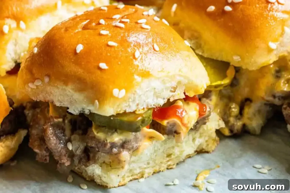 Close-up of a slider burger with sesame bun, melted cheese, beef patty, pickles, and ketchup on a parchment paper.